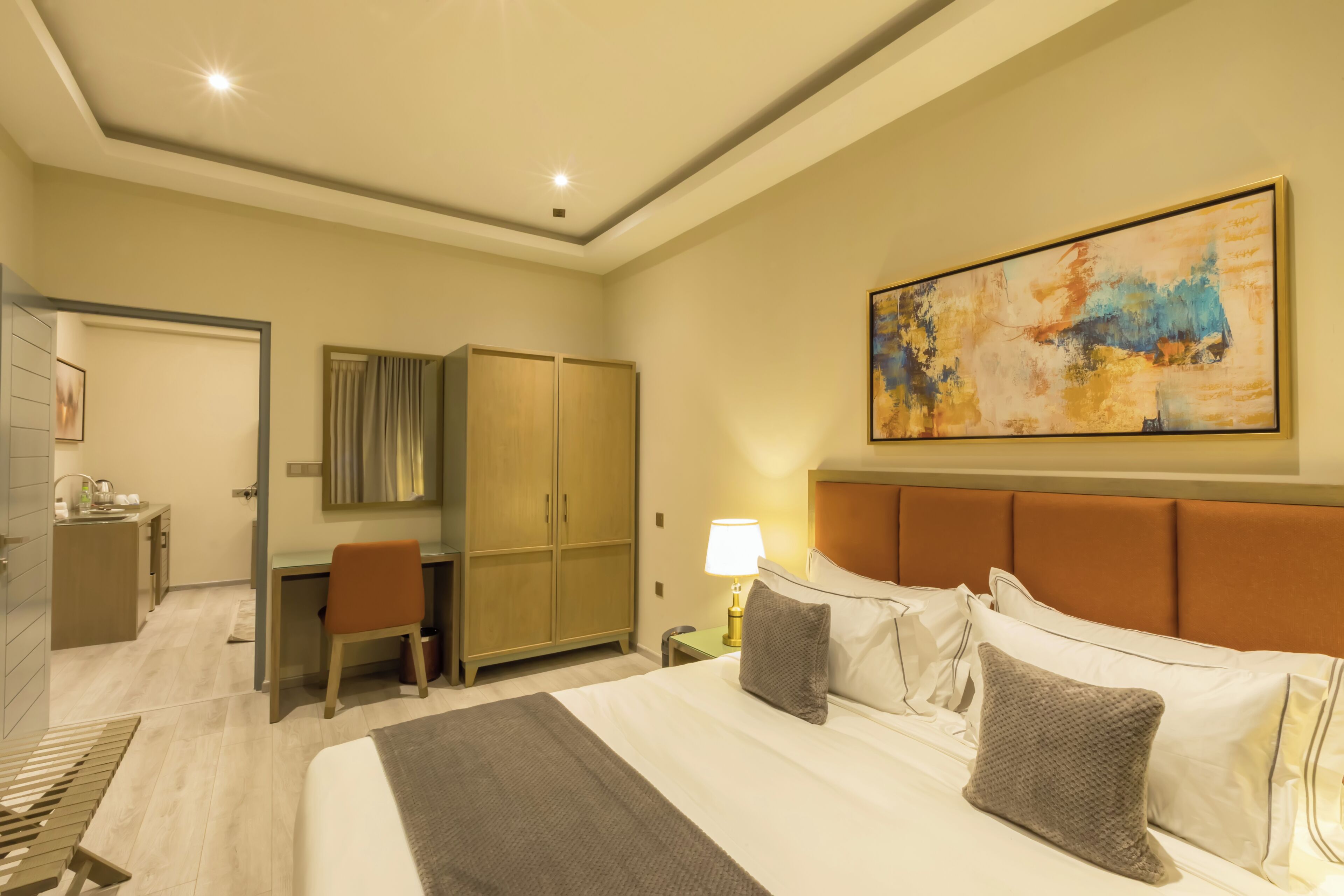 A hotel room with a bed, a desk, a wardrobe, a painting on the wall, and a nightstand with a lamp.