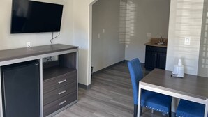Un espace de vie compact doté d’un téléviseur à écran plat fixé au mur, d’une petite kitchenette, d’un coin repas avec une table et des chaises bleues, et d’un meuble intégré avec des tiroirs.