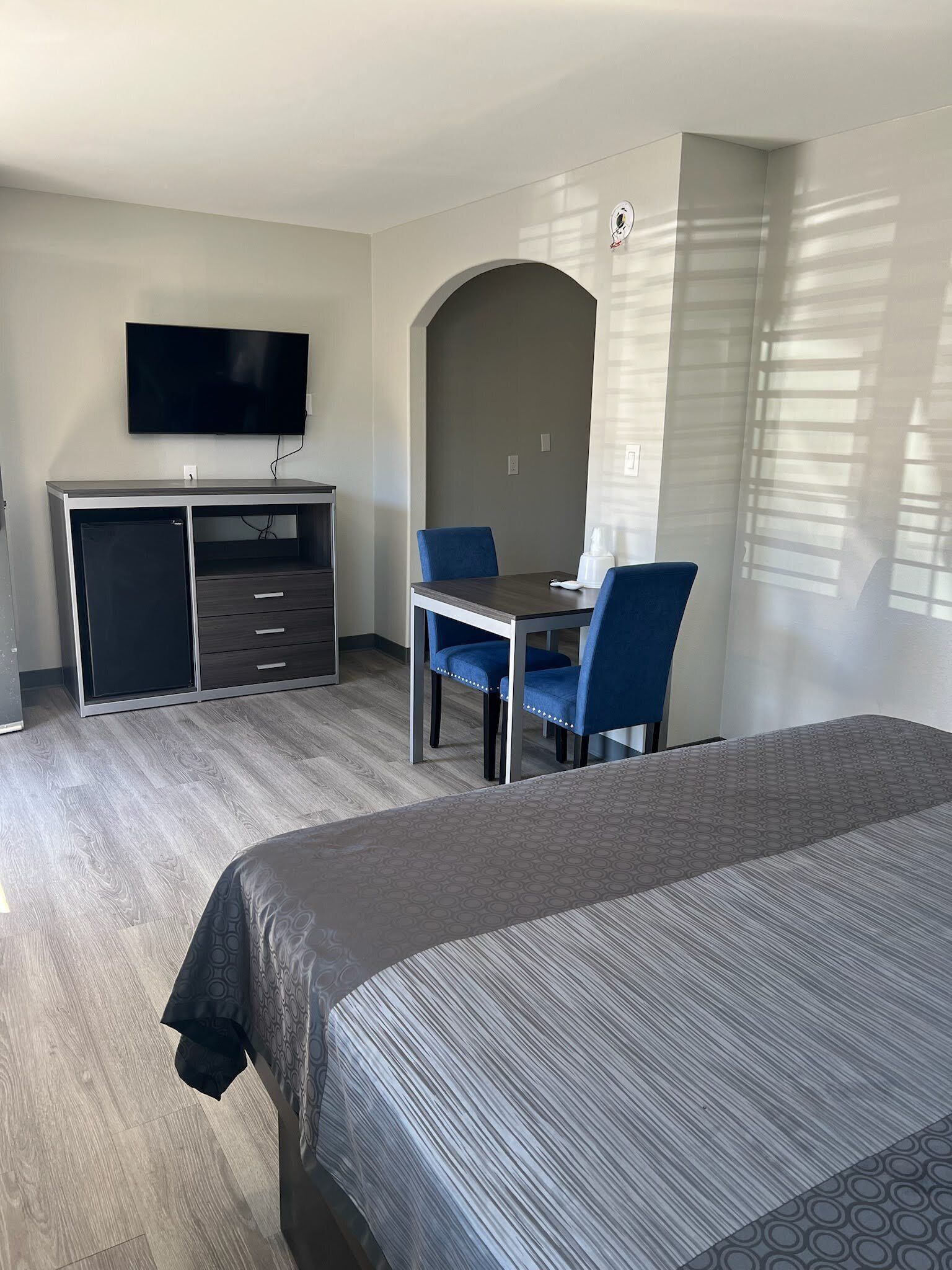 A hotel room with a bed, a desk with two chairs, a TV mounted on the wall, and a window with blinds.