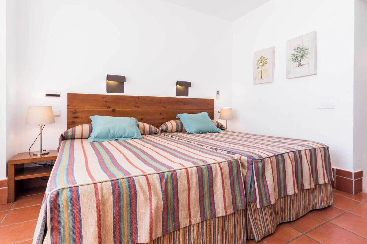 A bedroom with a wooden headboard, a striped bedspread, two pillows, a bedside table with a lamp, and a door to the right.