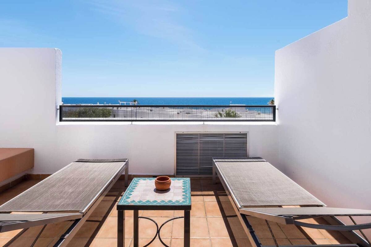 A balcony with a view of the sea, a table with a pot, and a bench.
