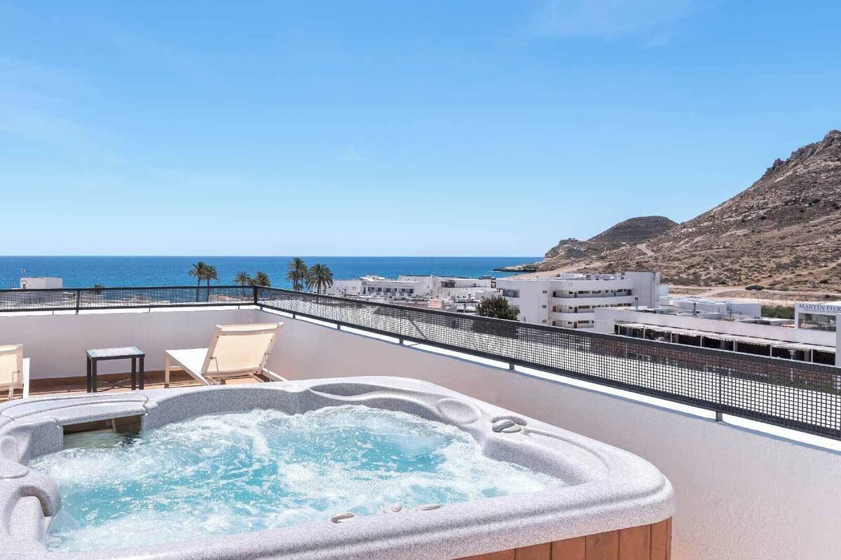A balcony with a hot tub, two chairs, and a view of the sea and buildings.