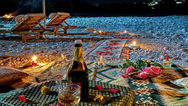 A beach setting with a table set for a meal, including a bottle, glasses, and flowers. - River Beach Resort - Trishuli (Dhussa)