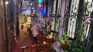 Um corredor externo estreito com cadeiras e mesas de madeira, plantas em vasos e luzes decorativas penduradas.