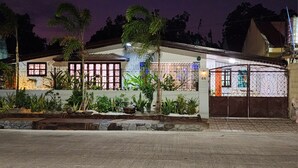 Uma casa de dois andares com portão fechado, cercada por um jardim paisagístico com diversas plantas e um pequeno espelho d'água.