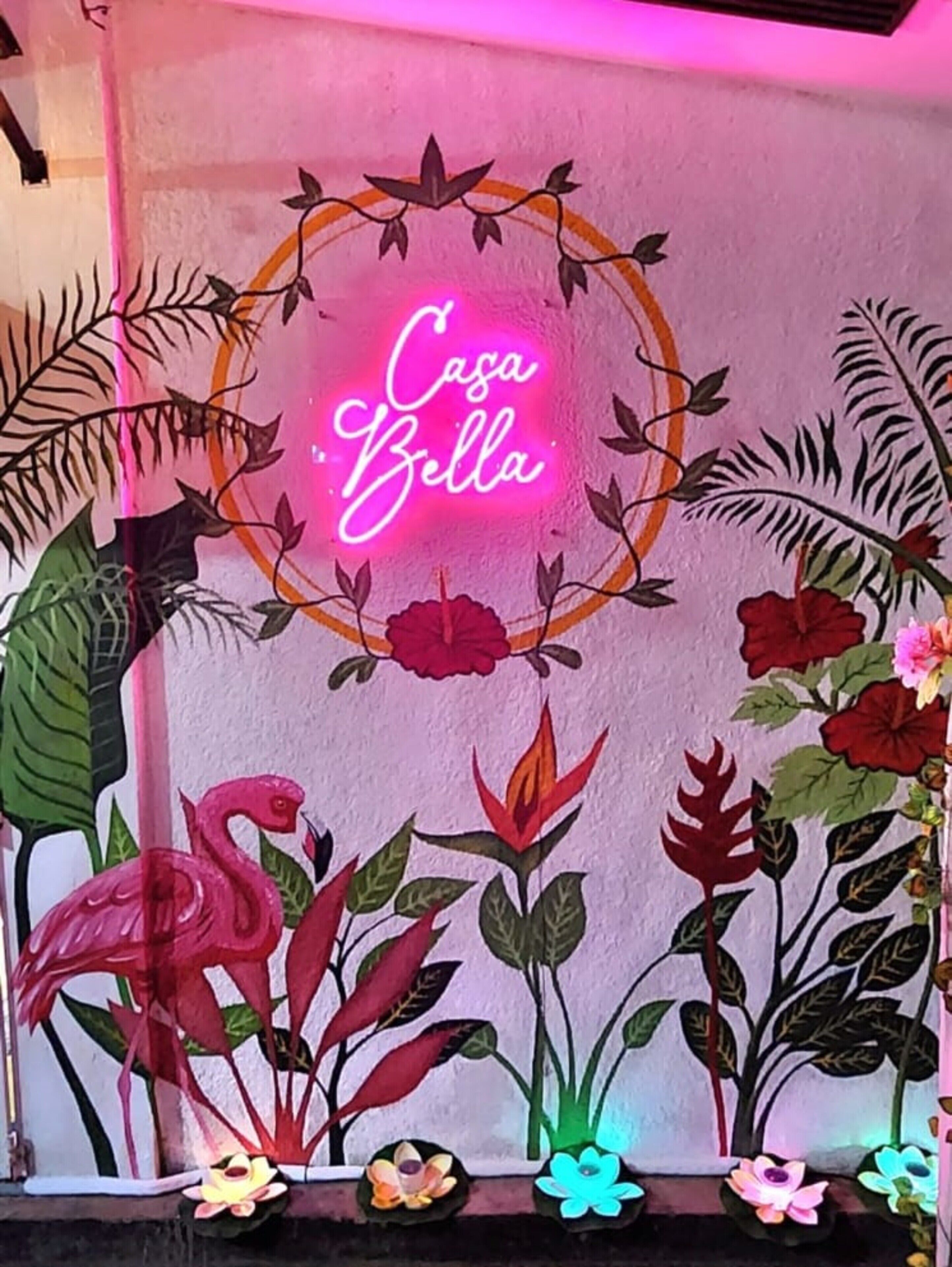 A wall with a neon sign that reads "Casa Bella" surrounded by floral and plant decorations.