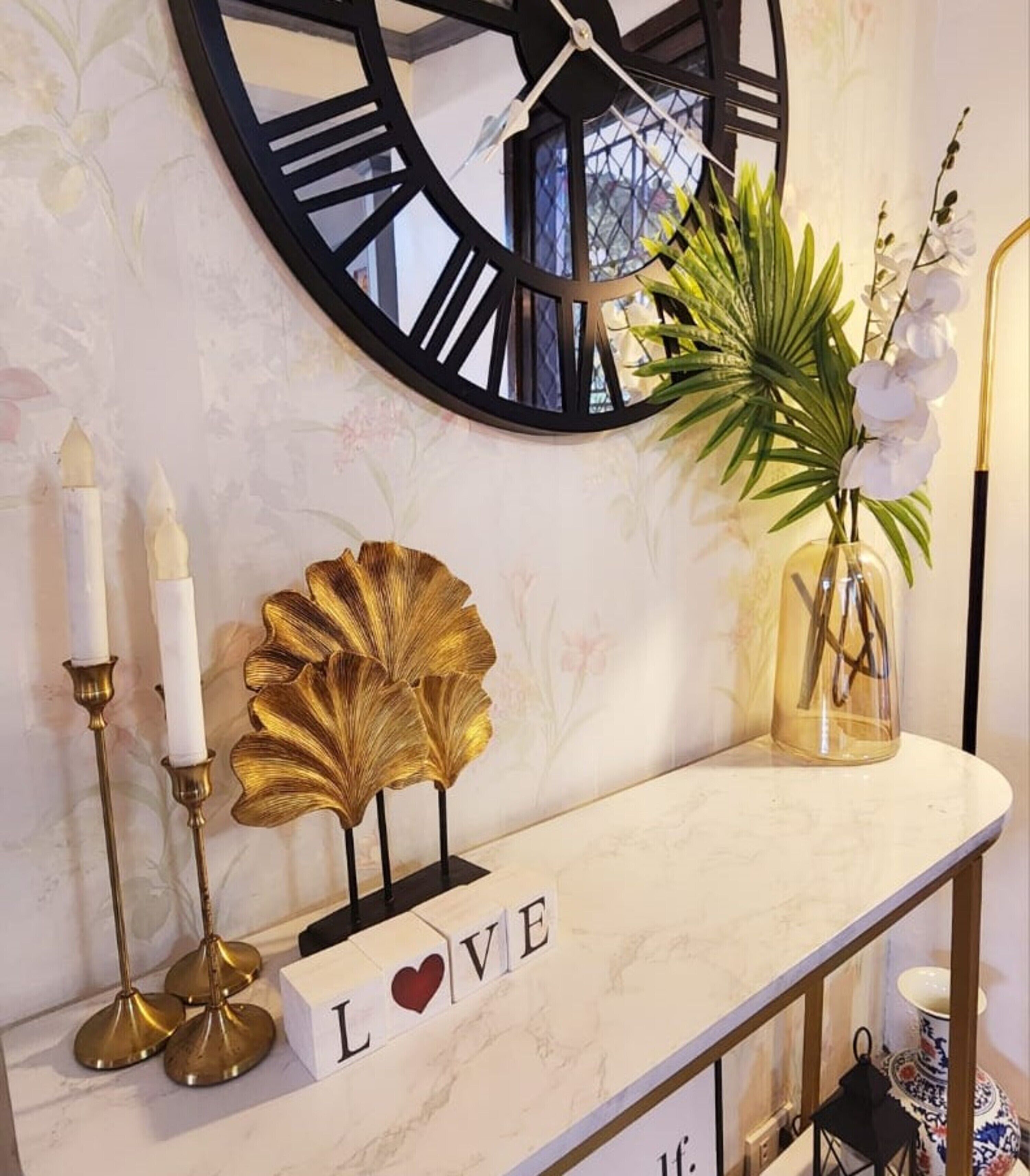 Ein Konsolentisch mit Marmorplatte, dekoriert mit Ziergegenständen, darunter eine große Uhr, Kerzen, eine Goldblatt-Skulptur, eine Orchideen-Vase und ein Block mit der Aufschrift „LOVE“.