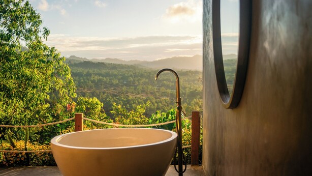 A large, round, beige sink with a curved faucet, positioned on a wooden floor with a view of a lush, green landscape through a window. - Alam Caldera Camp & Resto  (Kintamani)