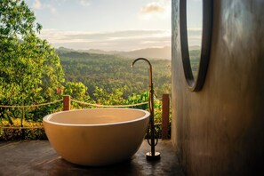 A large, round, beige sink with a curved faucet, positioned on a wooden floor with a view of a lush, green landscape through a window. - Alam Caldera Camp & Resto  (Kintamani)