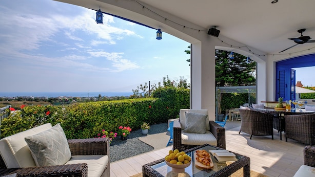 Un patio techado al aire libre con mobiliario de mimbre, una mesa de vidrio con fruta y vistas al mar.