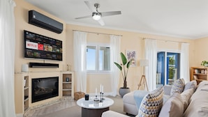 Un salón con televisor de pantalla plana, chimenea, ventilador de techo y un ventanal con cortinas.