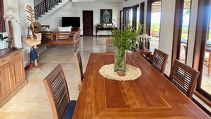 A spacious dining area with a wooden table and chairs, a staircase in the background, and a view of the outdoors through large windows.