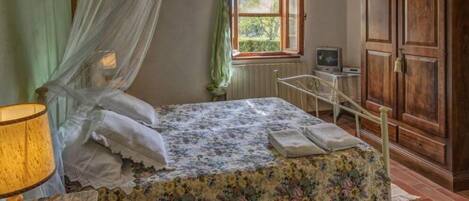 Une chambre à coucher avec un lit, une table de chevet avec une lampe, une armoire en bois, une fenêtre avec des rideaux et un radiateur.