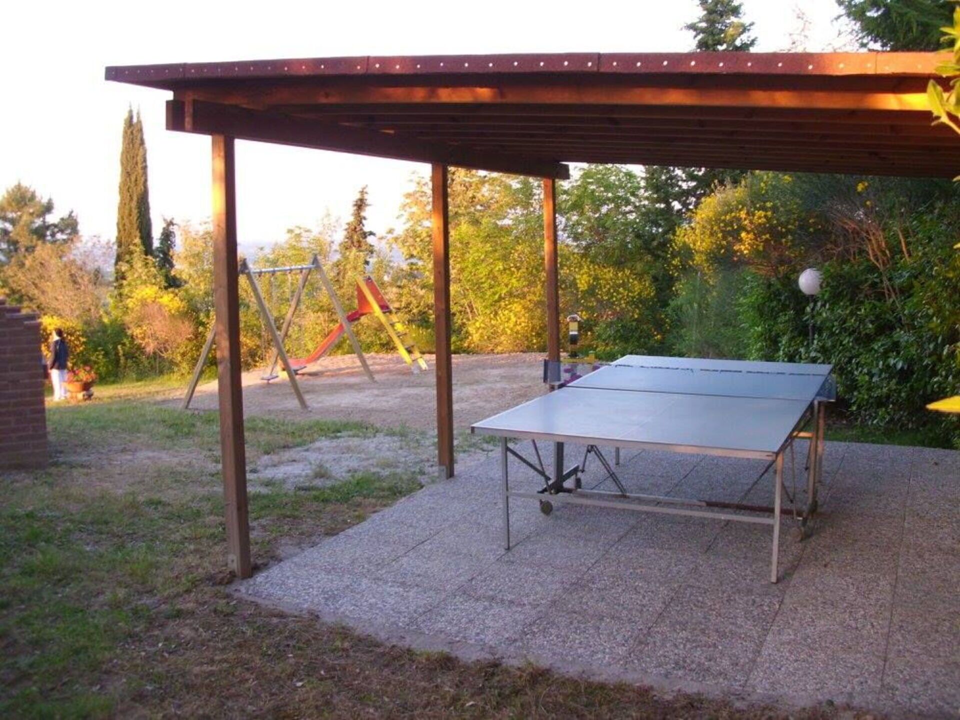 A ping pong table under a wooden pergola in a garden with a playground in the background.