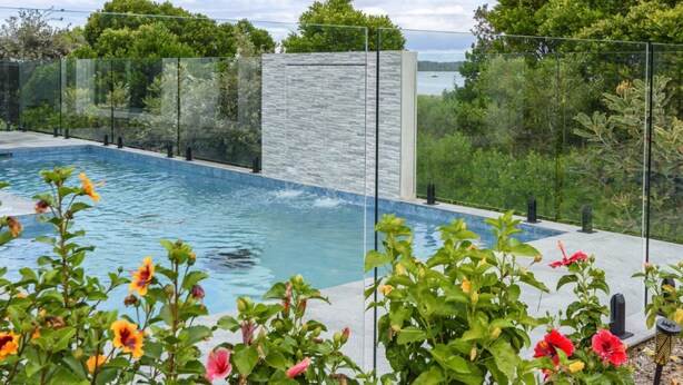 Una piscina con cerramiento de vidrio, rodeada de exuberante vegetación y un muro de piedra.