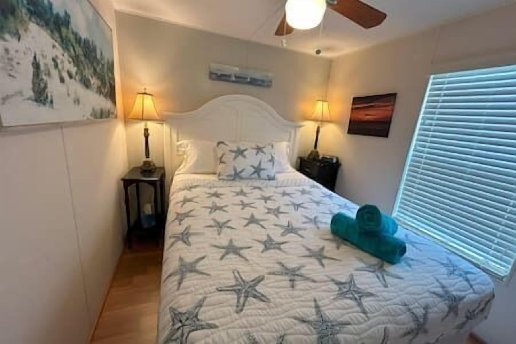 A bedroom with a bed, two bedside lamps, a ceiling fan, a painting on the wall, and a window with blinds.