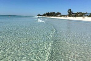 A beach with clear shallow waters, a sandy shore, and a few buildings in the background. - Waterfront with Unbelievable Views + Fabulous Lanai on the Water (Bradenton Beach)