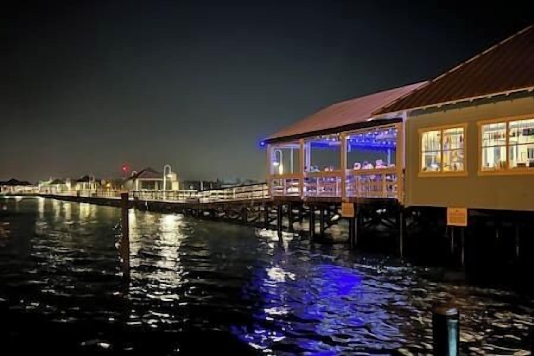 Un edificio frente al agua con un espacio exterior cubierto, un puente al fondo y una masa de agua que refleja luces.