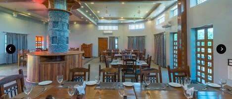 A spacious dining area with multiple tables and chairs, a central stone pillar, and large windows allowing natural light.