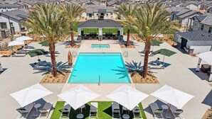 Une piscine entourée de chaises longues et de parasols, avec des palmiers et un bâtiment en arrière-plan.
