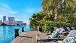 A poolside area with lounge chairs, a wooden deck, and a view of a city skyline across the water. - Spectacular Studio, Amazing amenities (Hallandale Beach)