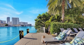 A waterfront pool area with lounge chairs, a palm tree, and a city skyline in the background.