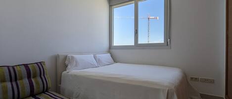A small, neatly arranged bedroom with a single bed, a striped sofa, and a window with a view of a construction crane.