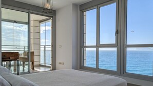 A bedroom with a large window offering a view of the sea, a balcony with a table and chairs, and a bed with a white bedspread.