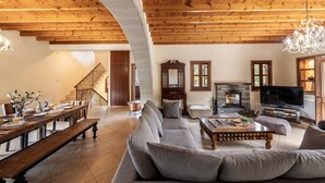 A spacious living room with a wooden ceiling, a fireplace, a television, and a dining area with a long table and benches.