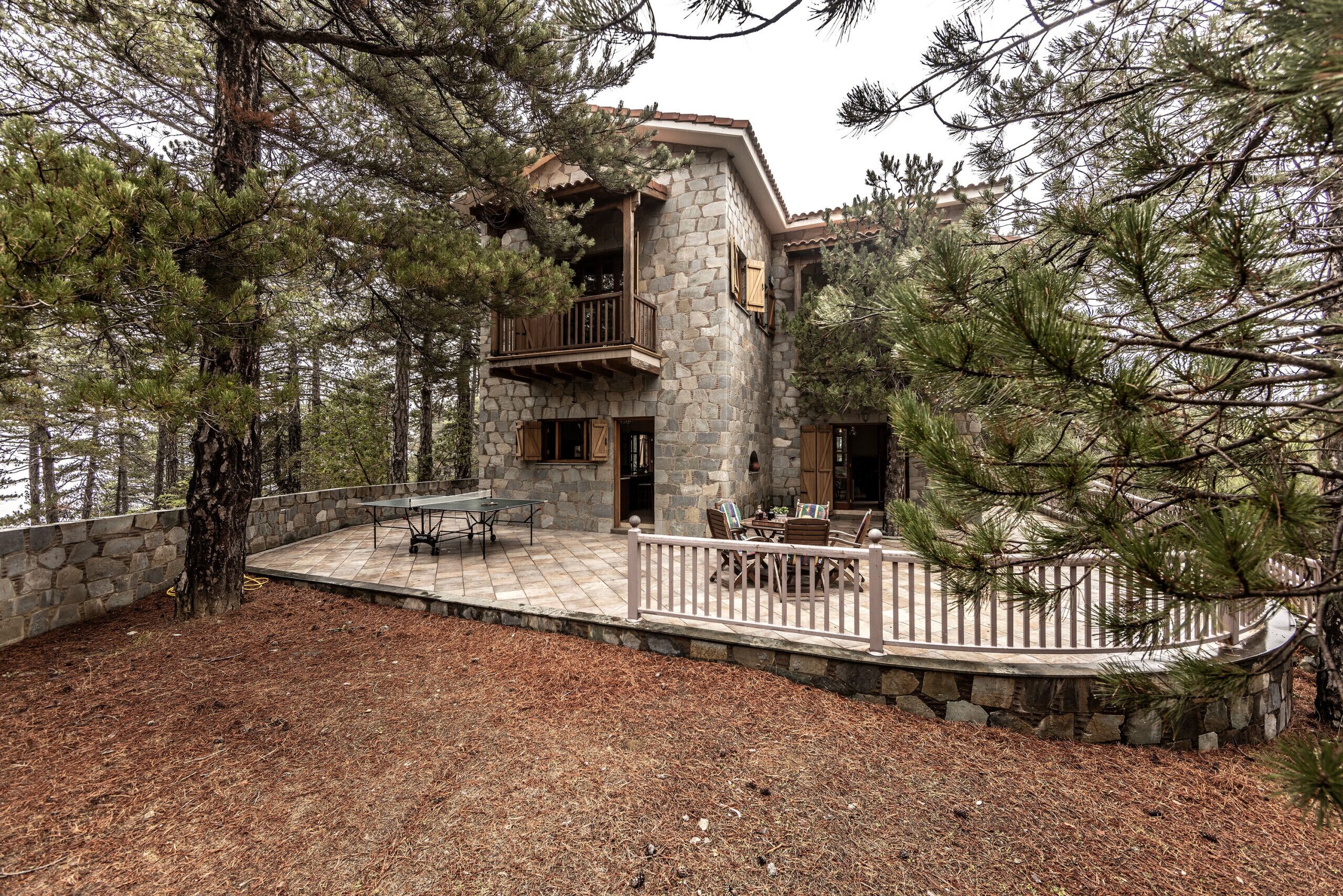 A two-story stone house with a balcony, surrounded by pine trees.