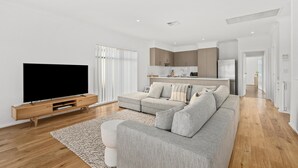 Una sala de estar moderna con un televisor de pantalla plana, un sofá seccional gris, piso de madera y una cocina al fondo.