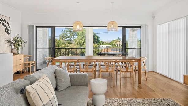 Un salón moderno con sofá, mesa de comedor y sillas, ventanas amplias con persianas y vistas a zonas verdes.