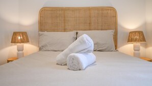 Una cama bien hecha con sábanas blancas y dos toallas enrolladas, con dos lámparas de noche de madera a cada lado.