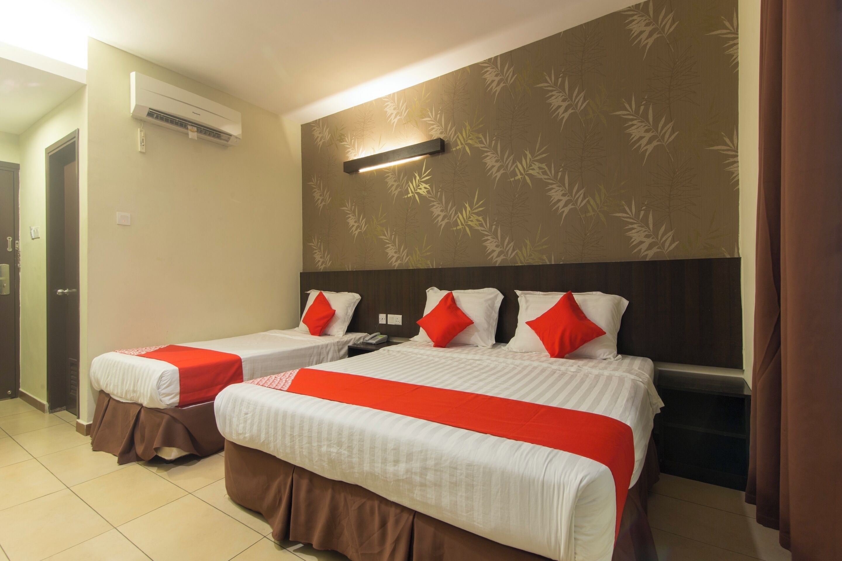 A hotel room with two beds, each with red and white bedding, a wall-mounted air conditioner, and a patterned wallpaper.