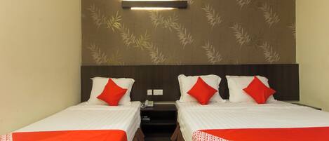 A hotel room with two beds, each with white bedding and red accents. There is a wall-mounted light fixture above the beds, and a small nightstand with a phone between them.