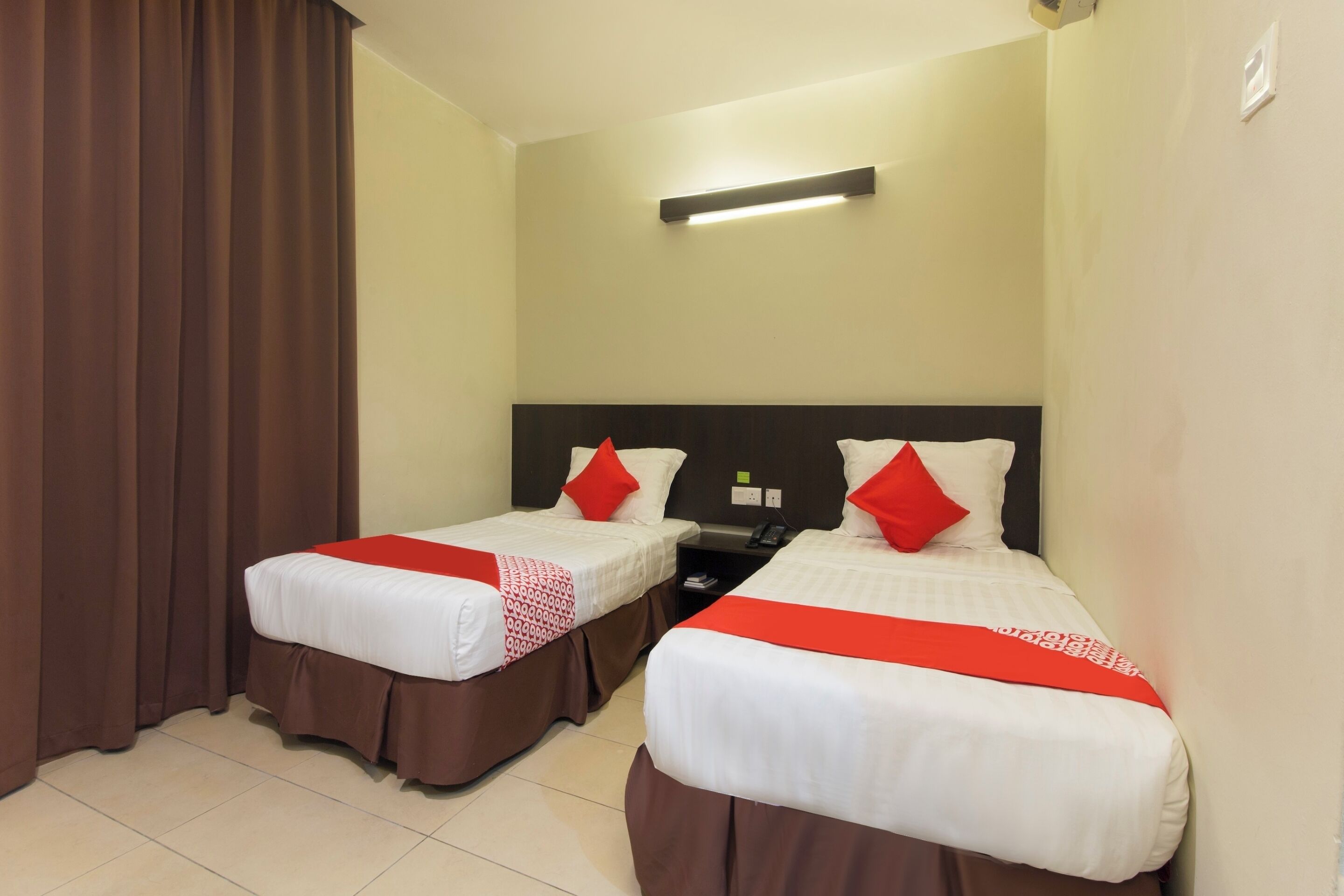 Une chambre d’hôtel avec deux lits, chacun doté de draps blancs et d’éléments décoratifs rouges, une table de chevet et un luminaire au plafond.