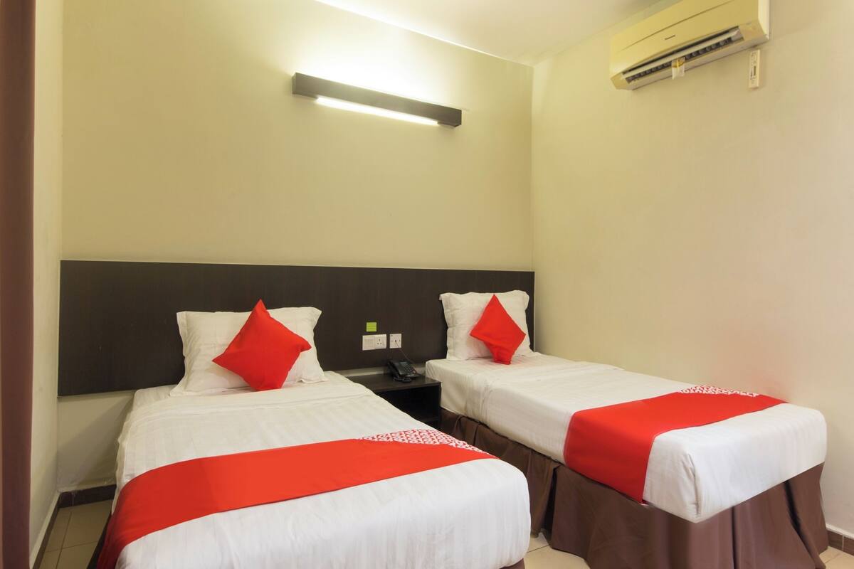 A hotel room with two beds, each with white bedding and red accents, a headboard, a bedside table, and a wall-mounted air conditioner.