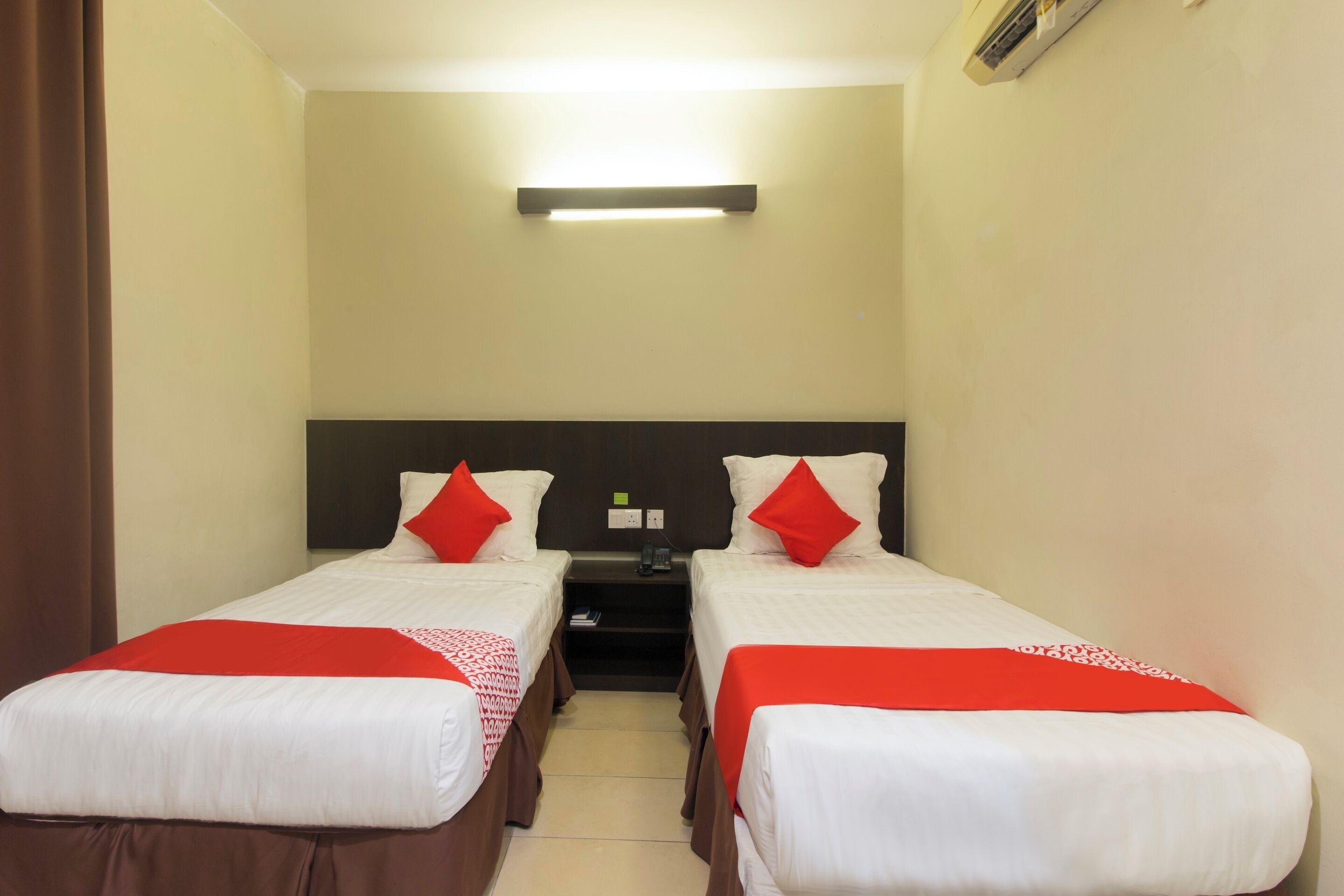 A hotel room with two single beds, each with a red and white bedspread, a nightstand, and a wall-mounted air conditioning unit.