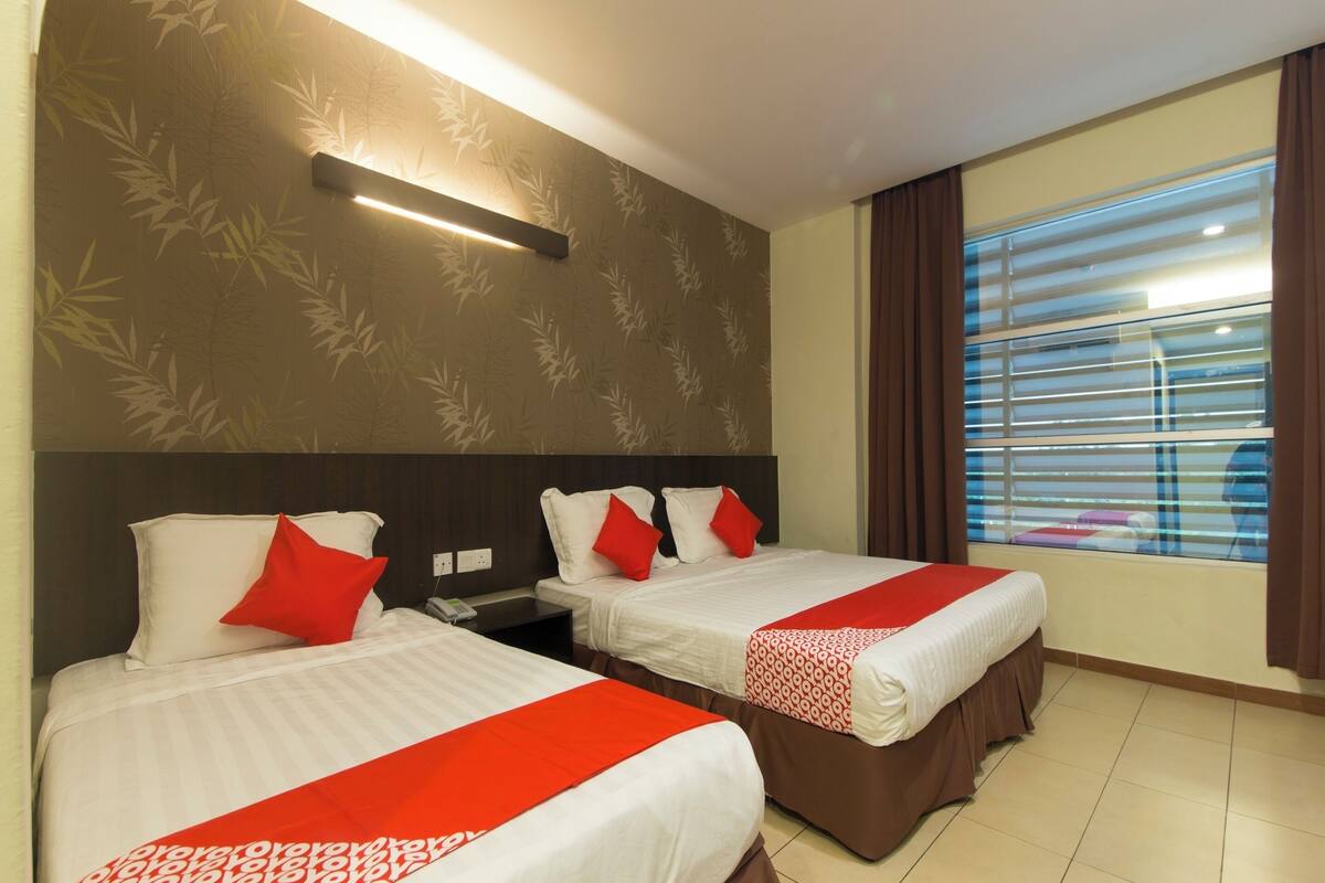A hotel room with two beds, each with white bedding and red accents, a nightstand, a wall-mounted lamp, and a window with blinds.