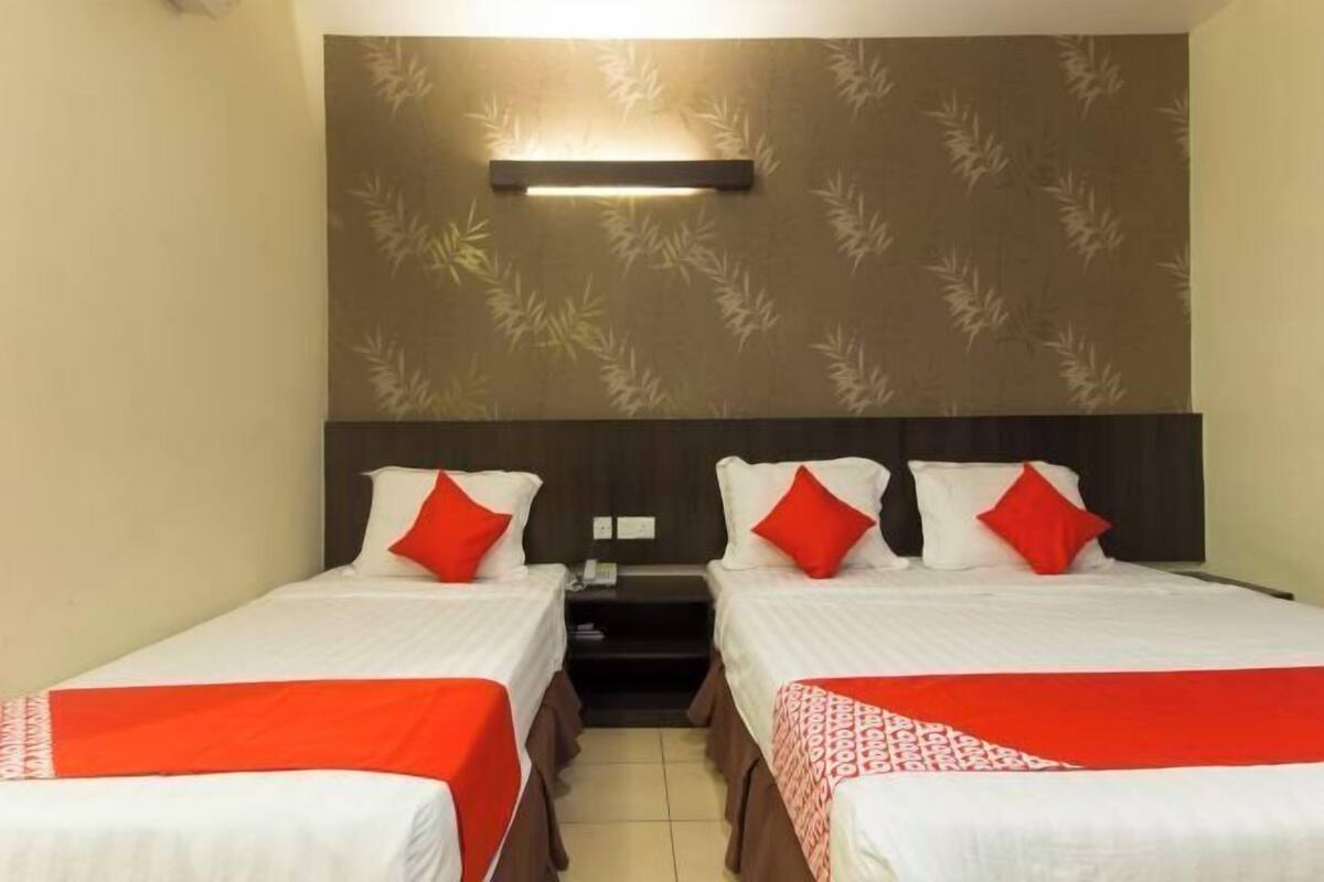 A hotel room with two beds, each with white bedding and red accents, a nightstand, and a wall-mounted light fixture.