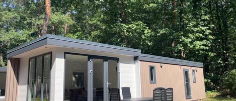 Une cabane moderne, de plain-pied, avec une entrée dotée d’une porte en verre, une terrasse avec une table et des chaises, et une vue sur une zone boisée.