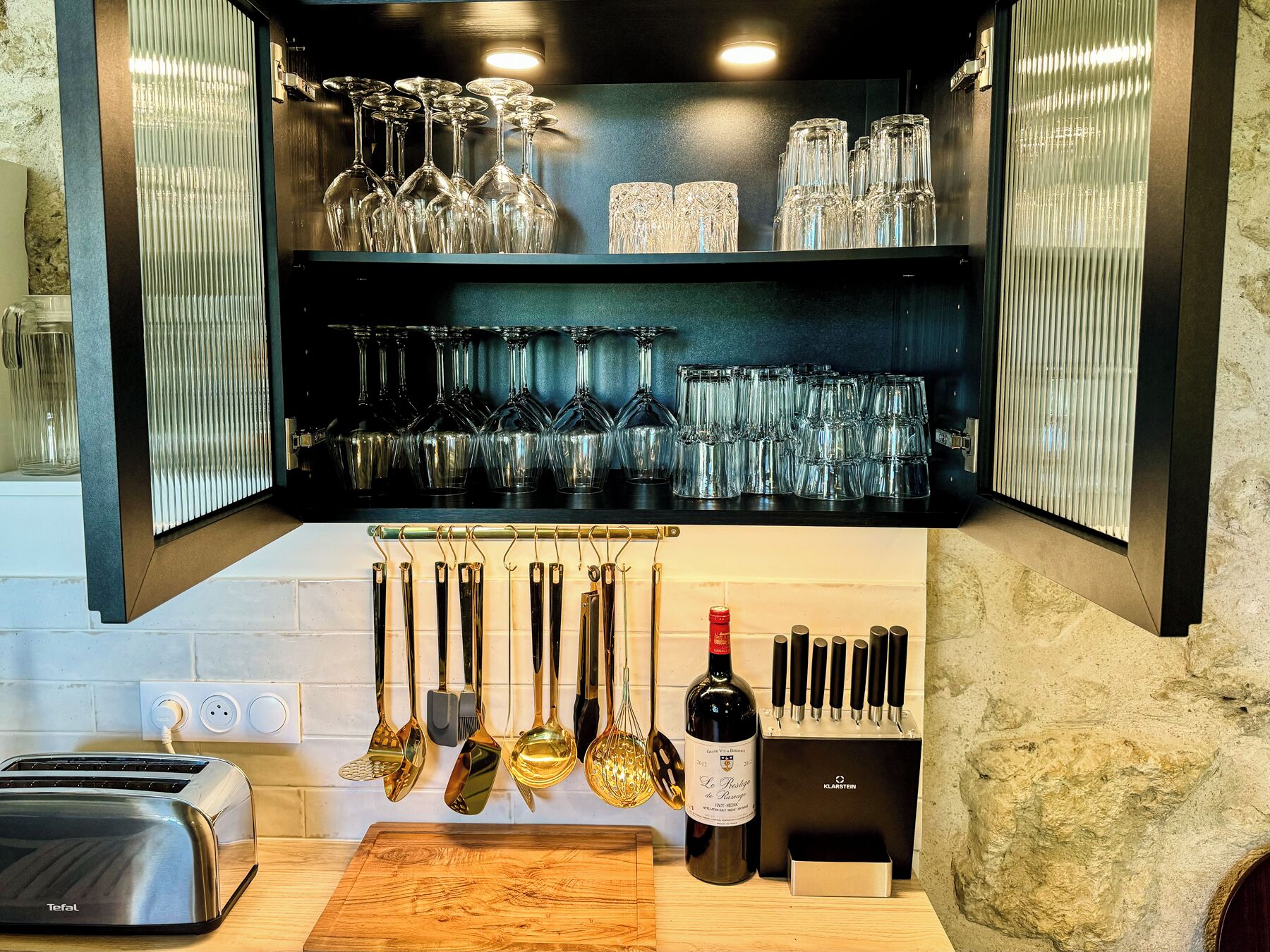 A kitchen with a built-in wine rack holding various glassware, a toaster, and hanging kitchen utensils.