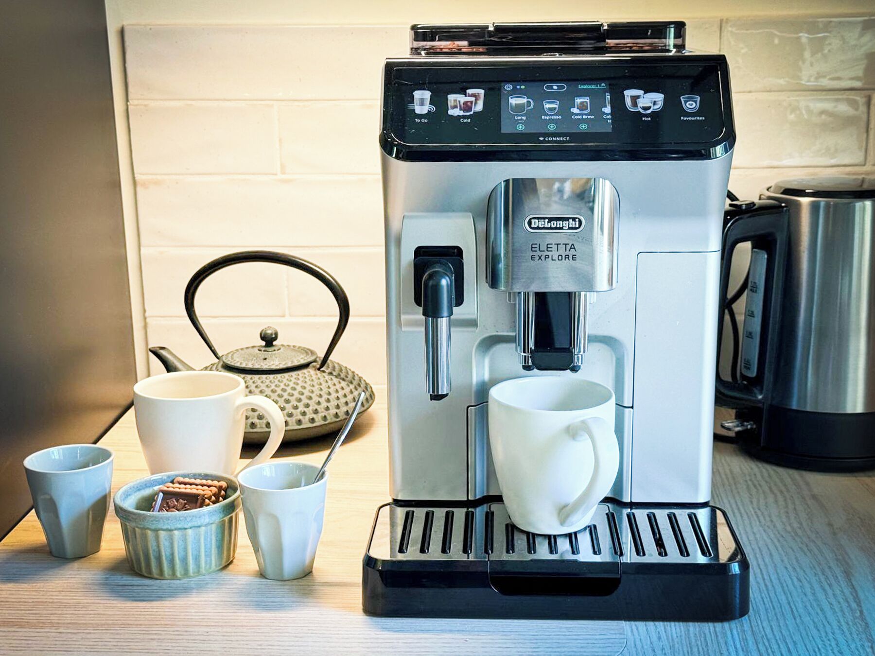 Una cafetera Delonghi Eletta Explore con pantalla digital, una taza de café, un hervidor de tetera y una tetera sobre una encimera de cocina.