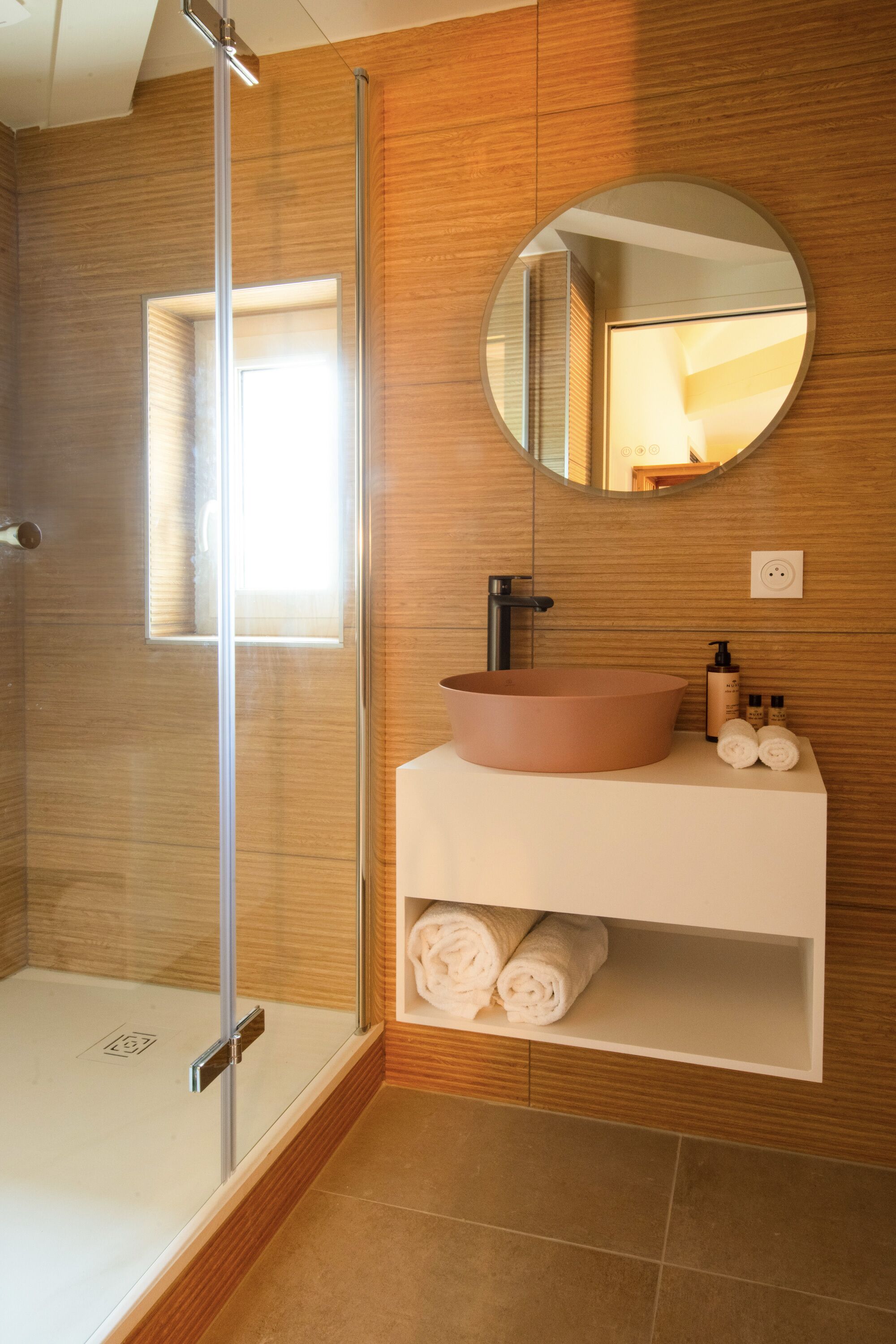 A modern bathroom with a glass-enclosed shower, a round mirror, and a white vanity with toiletries.