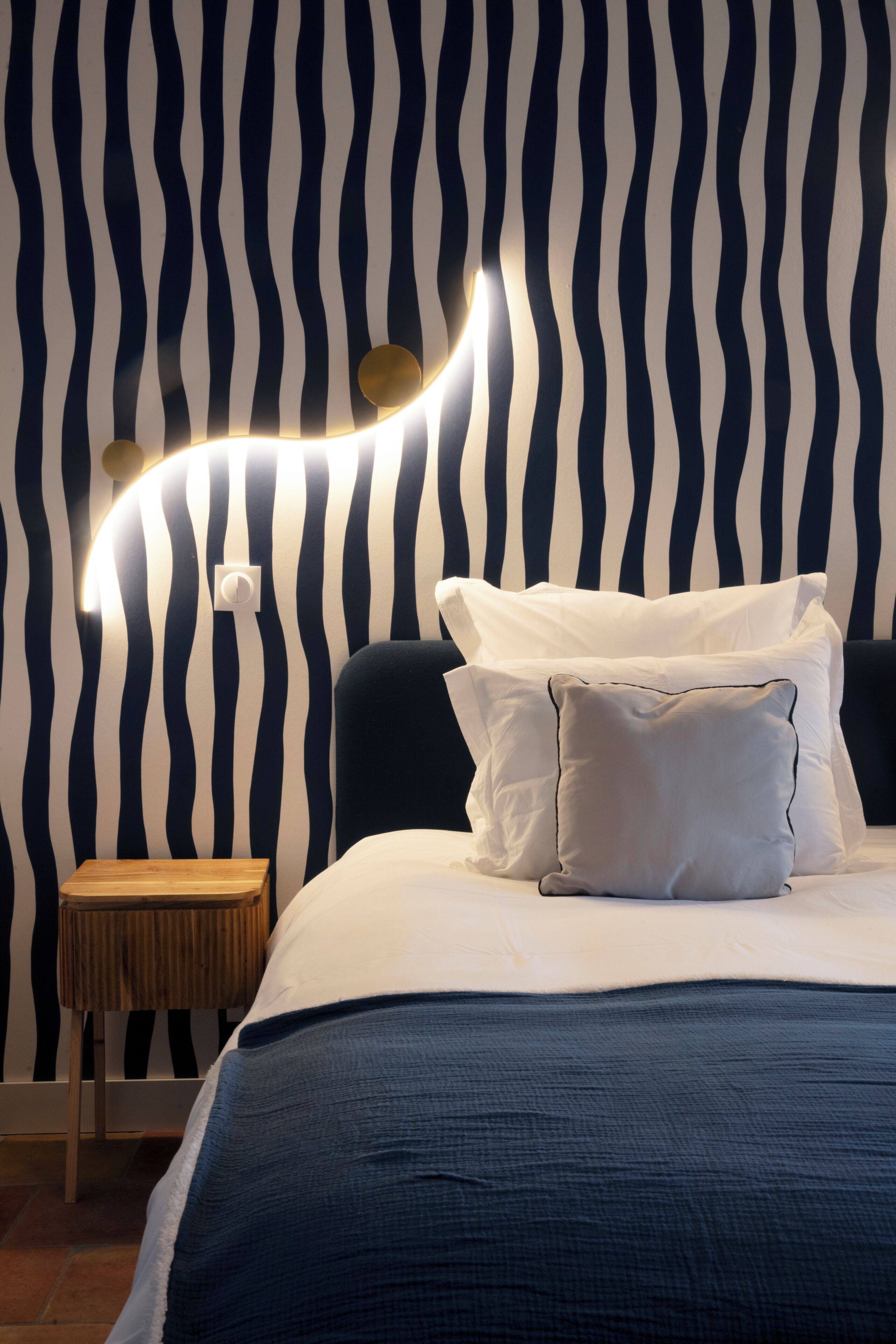A bedroom with a striped wall, a bedside table, and a bed with pillows.