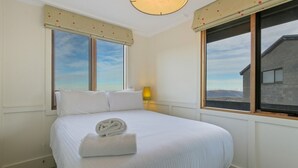 A neatly made bed with a rolled towel, two windows with curtains, a lamp, and a view of a building through the window. - Shamrock 1 - Mt Hotham (Hotham Heights)