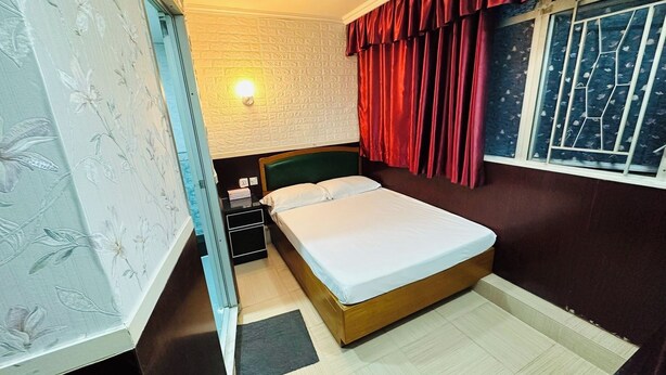 A room with a bed, a nightstand, a wall-mounted lamp, and a window with curtains. - Tai Tung Hostel (Kowloon)