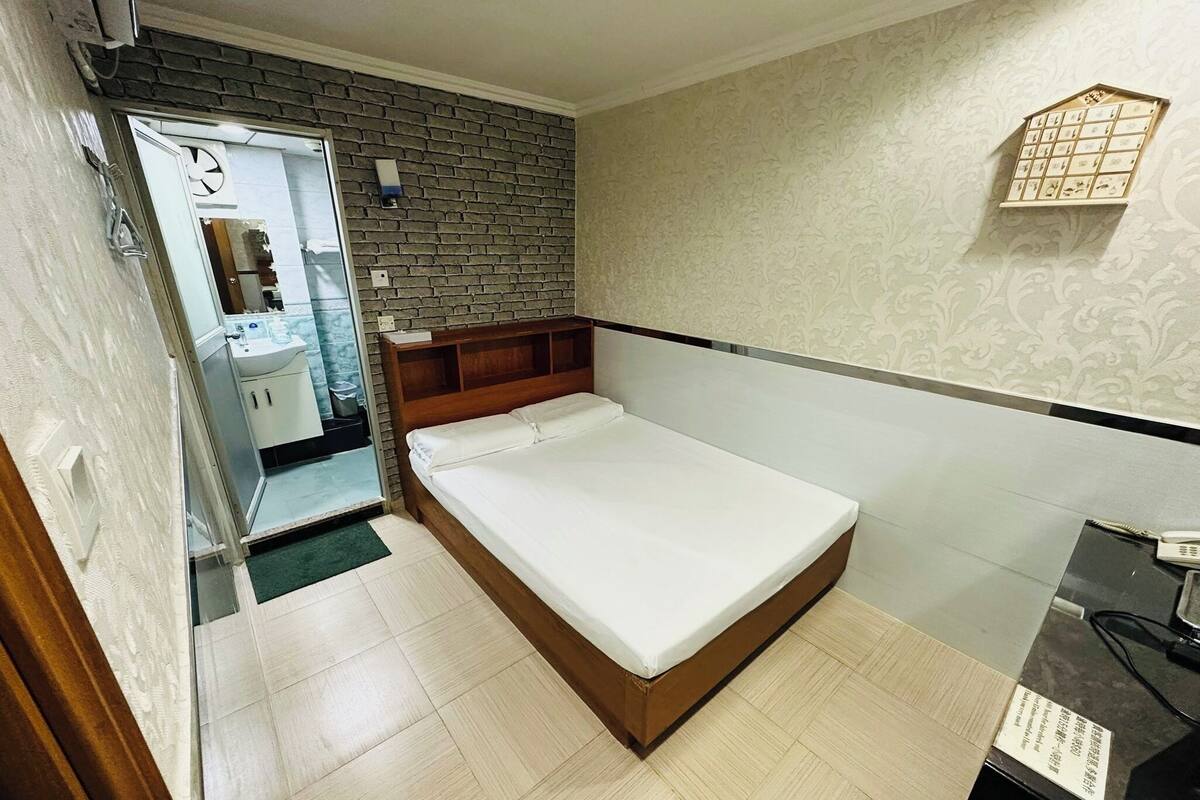 A bedroom with a wooden bed, a small bedside table, a mirror, and a bathroom visible through an open door.