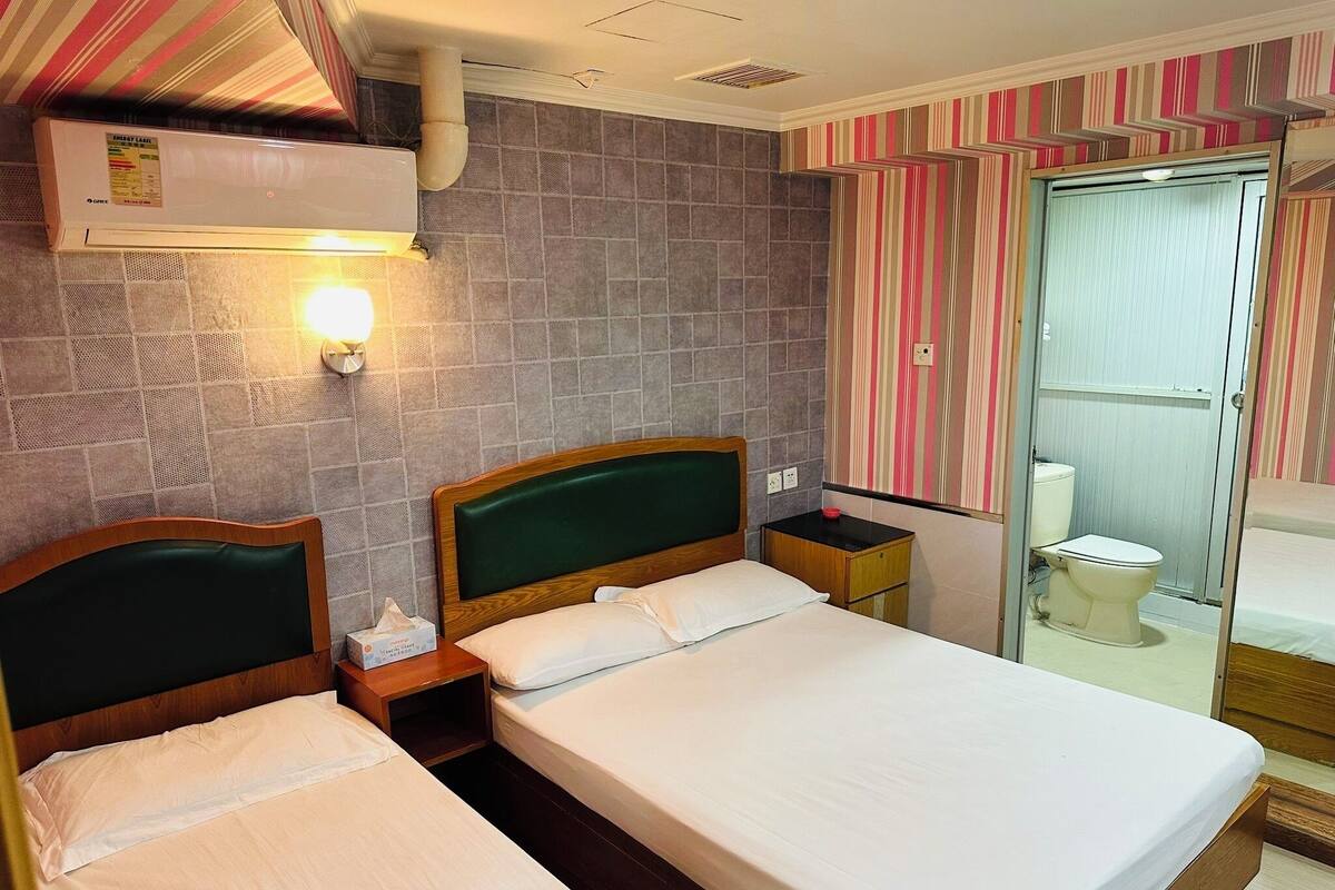 A hotel room with two beds, a bathroom with a toilet and a shower, and a wall-mounted air conditioner.