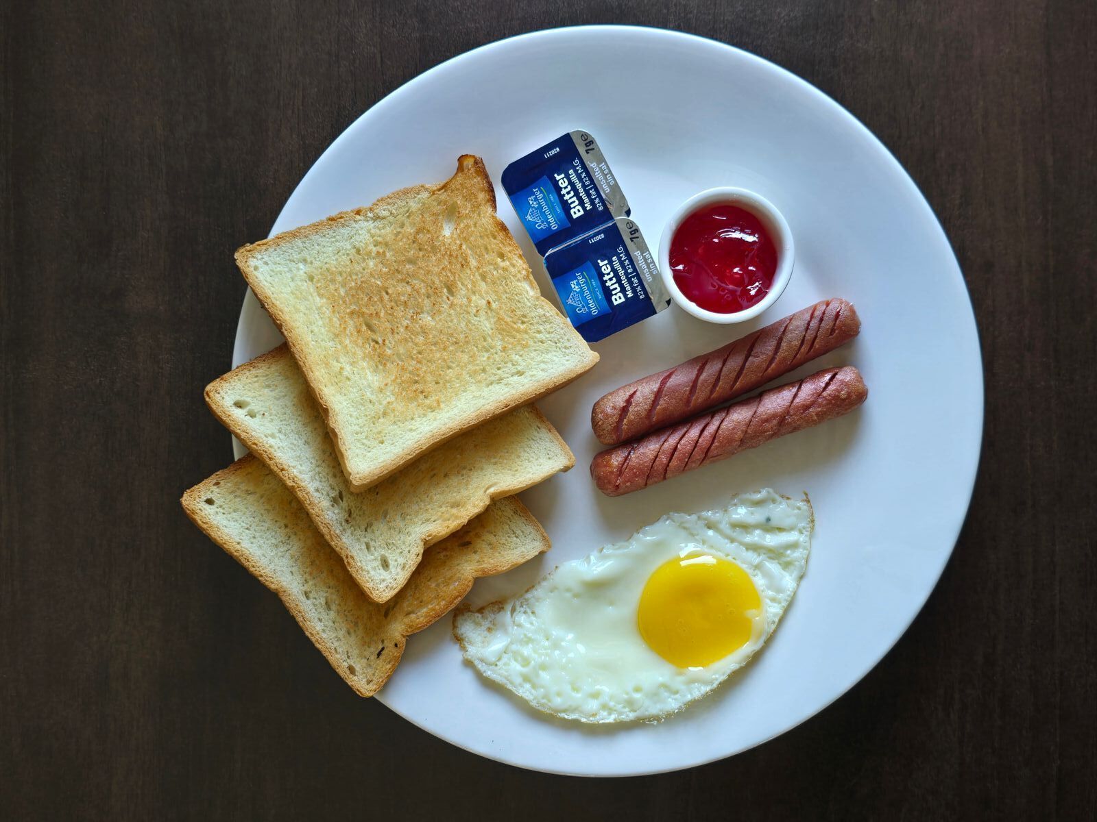 A plate with toast, sausages, a fried egg, butter, and ketchup.